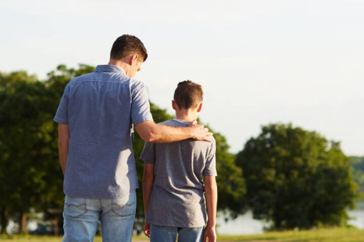 Man with arm around a boy's shoulder standing on a grassy field by a lake.