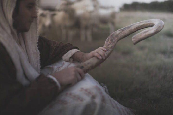 Person in a hooded shawl sitting in a field, holding a large curved ram horn (shofar).