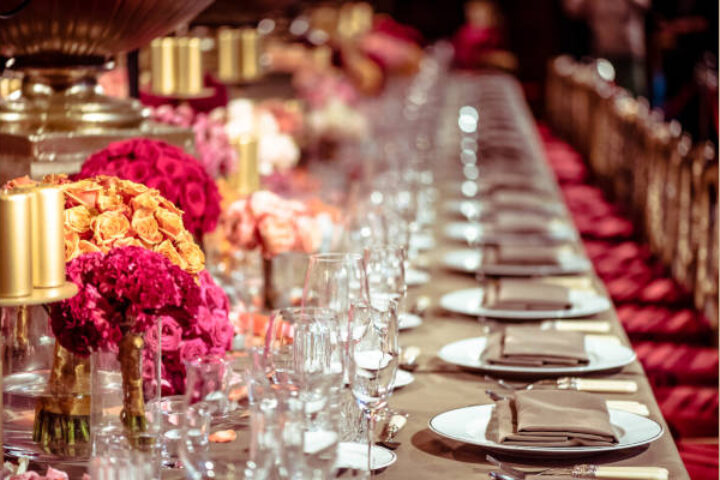 Long banquet table set for a formal dinner with pink and orange flower centerpieces, glassware, and folded napkins along the plates.
