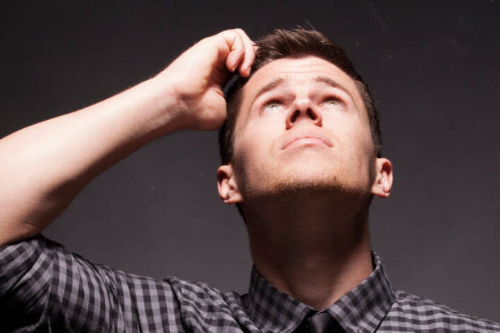 Man in a checkered shirt looks up with his hand on his head, appearing thoughtful against a dark background.